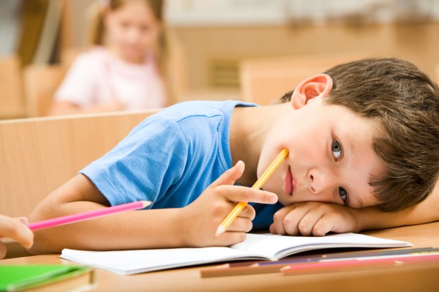 image of elementary age boy sitting at desk, leaning on arms with notebook under arm, pencil in hand.