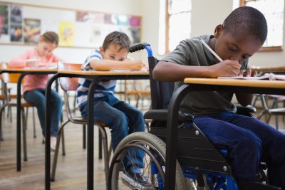 Children in a row at their desks in class; the first boy is in a wheelchair.