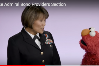 A woman in a Navy dress uniform is facing Elmo, the shaggy red Muppet from Sesame Street. They appear to be speaking to each other